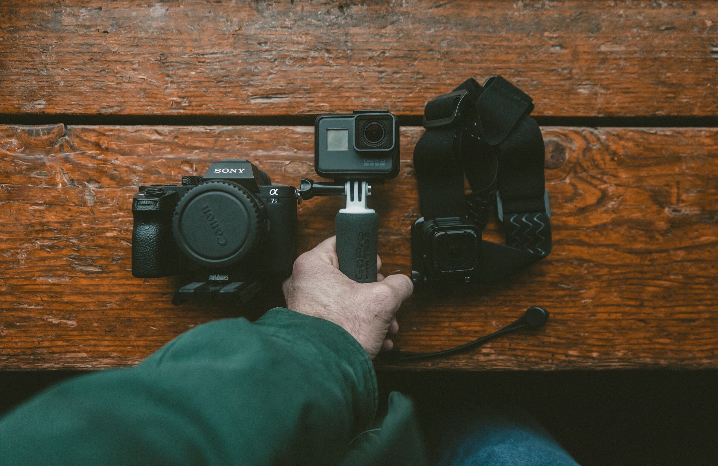 A hand holding an action camera beside a mirrorless camera on a wooden table